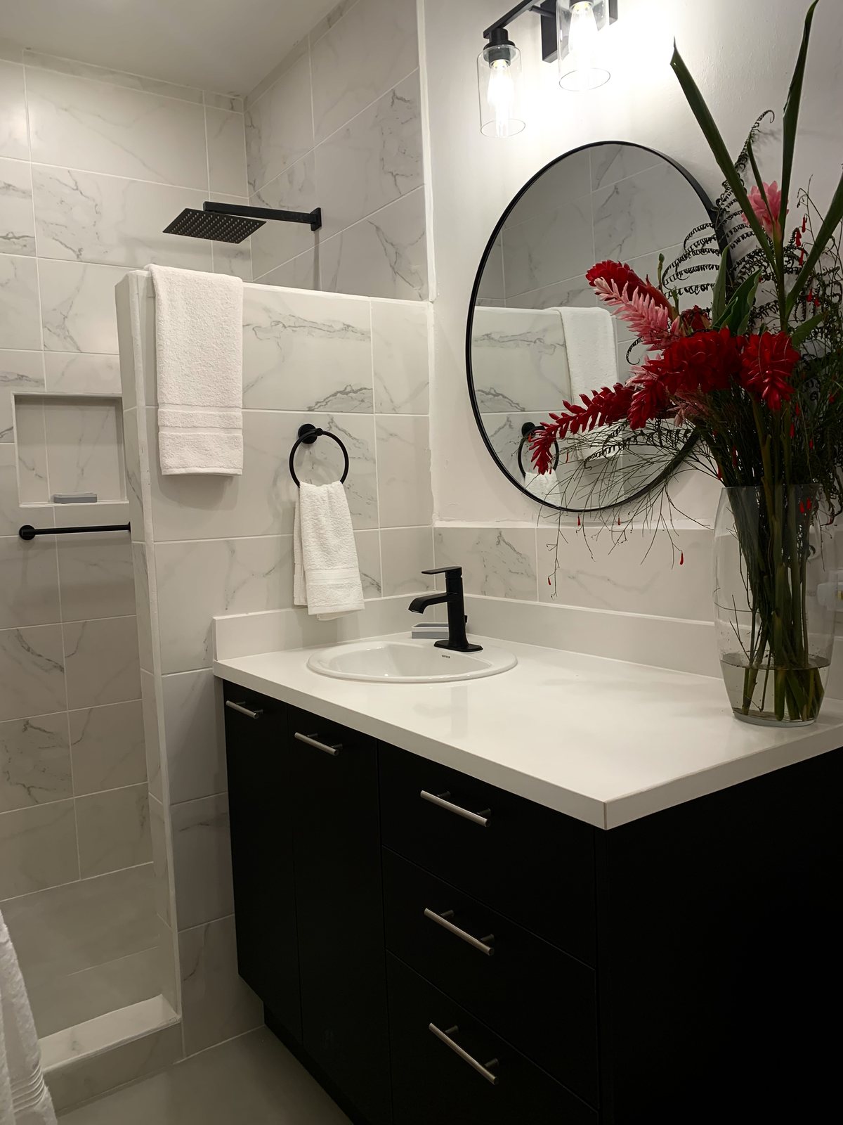 Bathroom with a black vanity, round black mirror, walk-in shower and red ginger flowers